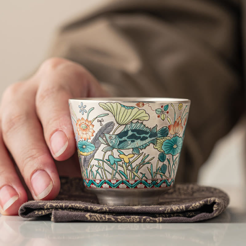 Fish Lotus Tea Cup – mandarin fish and lotus petals held on dark brocade, lifestyle shot, ceramic teacup