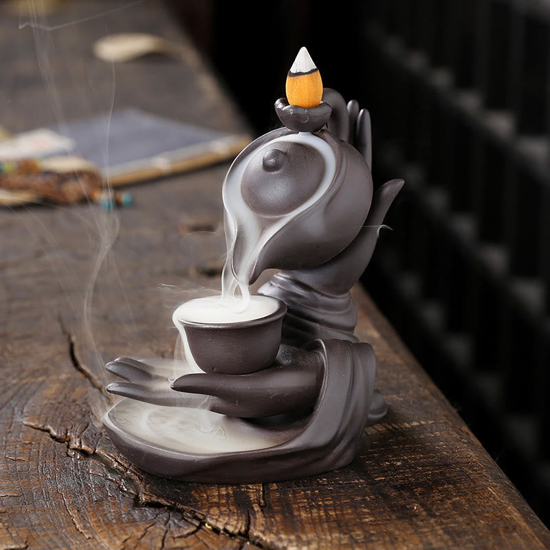 Buddha Hand Teapot Incense Burner – Stylized hands cradle cup, smoke cascading like waterfall, dark ceramic on wood