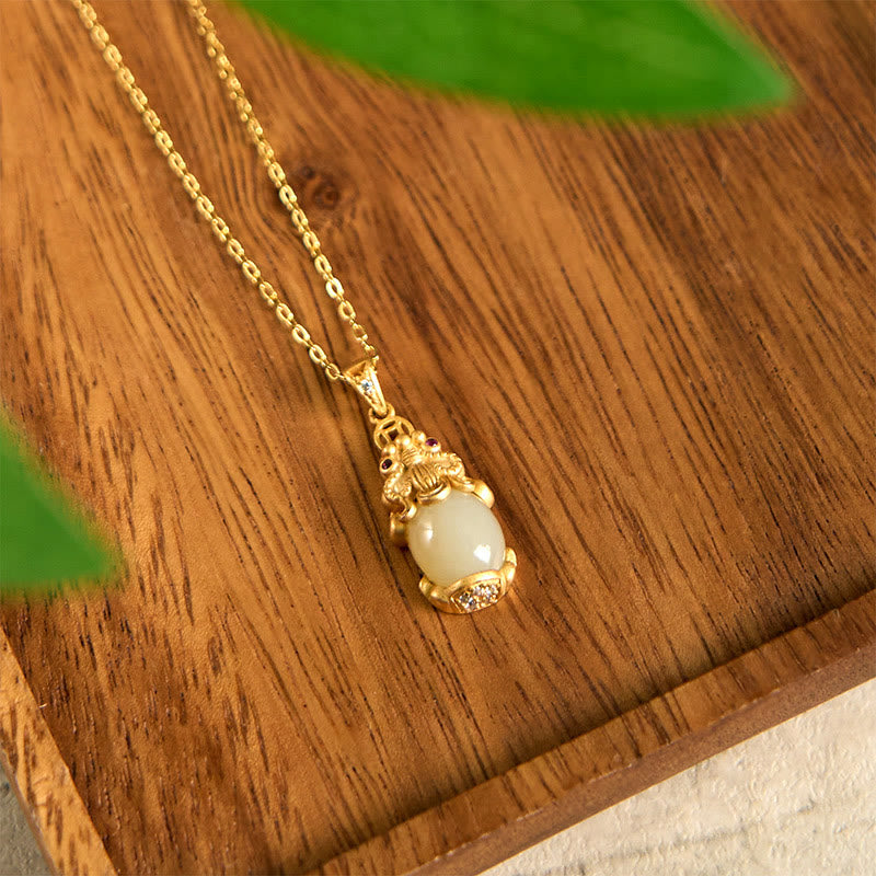 White Jade Pixiu Wealth Luck Pendant Necklace – gold chain jade pendant on warm wood surface with green leaves, good luck jewelry