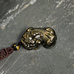 Gold Sheen Obsidian PiXiu Wealth Rope Necklace Pendant – flat lay side angle on gray stone, carved gold sheen PiXiu detail