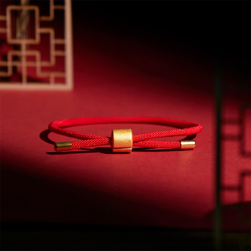 Braided red cord bracelet with matte gold cylindrical slider bead, dramatic studio lighting on crimson surface with Chinese lattice backdrop