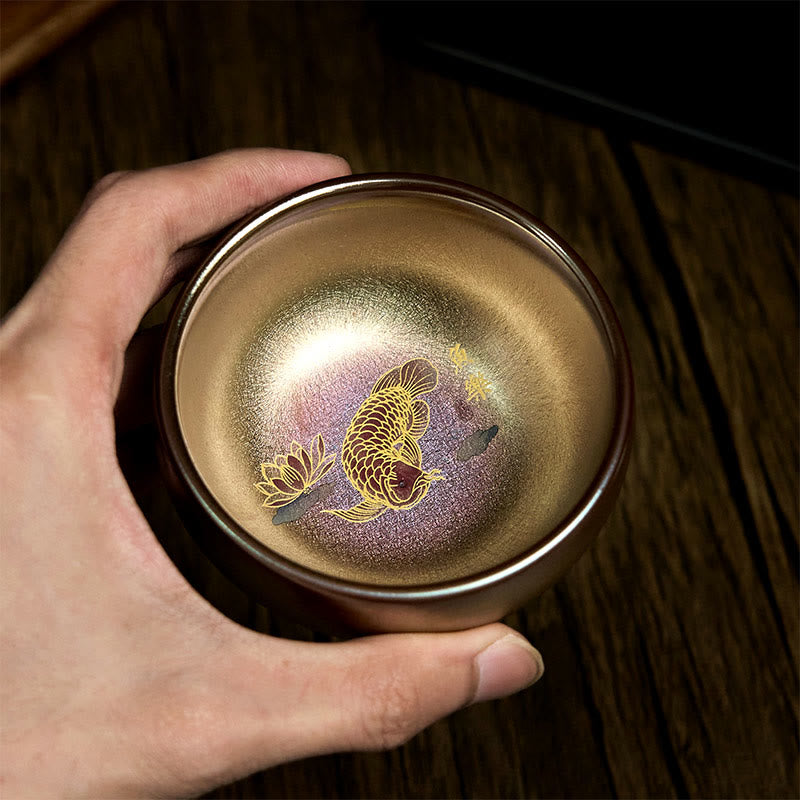Jianzhan Koi Fish Lotus Teacup – handheld overhead, iridescent gold-purple cup with koi and lotus, held in hand, dark wood