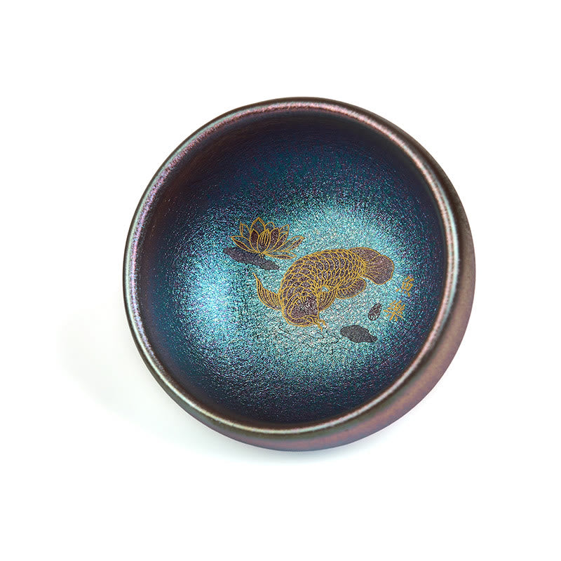 Jianzhan Koi Fish Lotus Teacup – handheld side view, iridescent bronze cup with gold koi, dark wood tea table setting