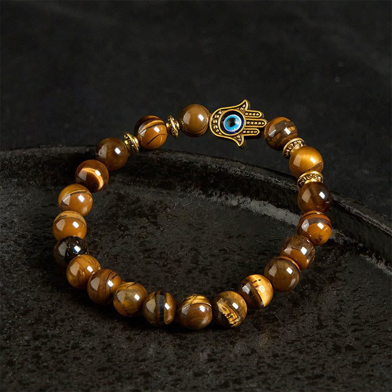 Tiger eye bracelet with gold evil eye Hamsa charm worn on wrist, black background