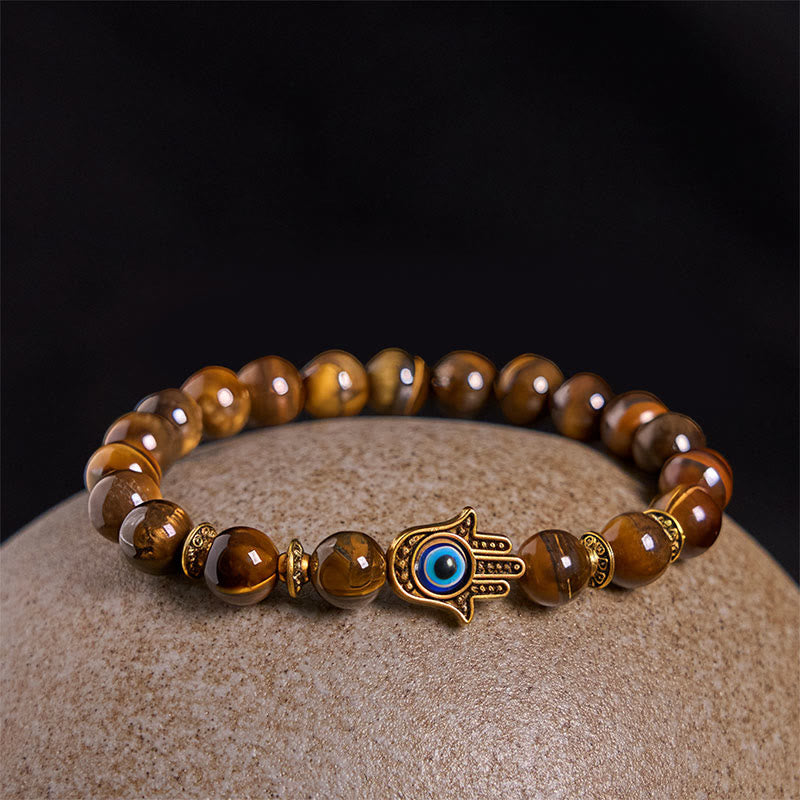 Tiger eye bead bracelet with gold evil eye Hamsa charm on beige stone surface, black backdrop