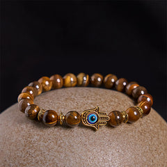 Tiger eye bead bracelet with gold evil eye Hamsa charm on beige stone surface, black backdrop