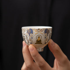 White Thangka Buddha Kung Fu Tea Cup – two hands holding front view, gold Buddha figure with blue auspicious clouds, serene