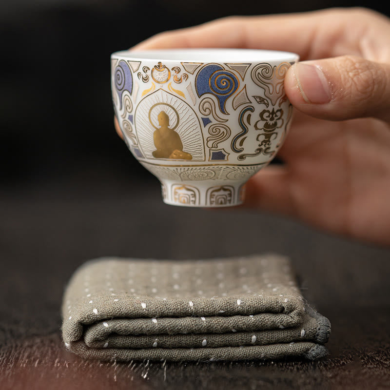 White Thangka Buddha Kung Fu Tea Cup – front view held above grey linen napkin, gold Buddha with blue cloud details, elegant