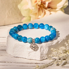 Apatite Lotus Silver Bracelet – on stone pedestal with cream flowers, warm light on blue apatite and silver lotus charm