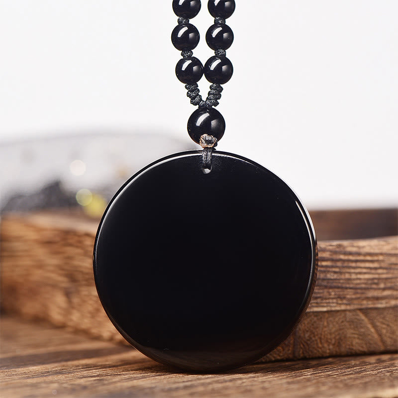 Black Obsidian Dragon Necklace - polished black disc pendant with beaded cord on wood surface, Strength