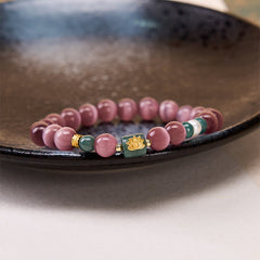 Purple Cat’s Eye Lotus Enlightenment Bracelet – Resting on dark ceramic dish, mauve cat’s eye beads with green jade lotus charm, mindful jewelry