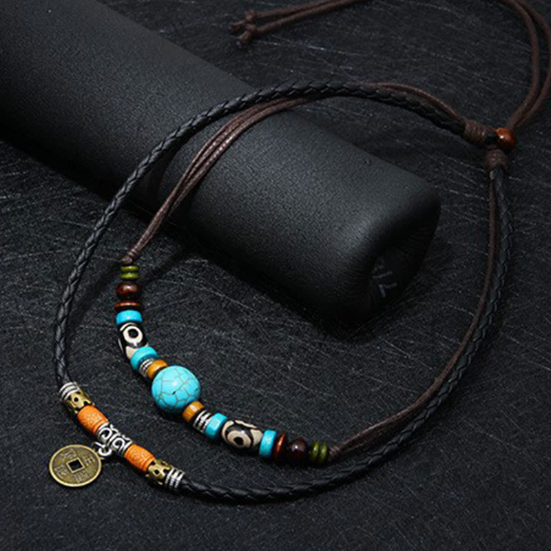 Turquoise Dzi Bead Protection Necklace – three-quarter angle flat-lay on dark textured surface showing turquoise bead centerpiece with matrix veining, Dzi pattern beads, amber accents, silver skull beads, and bronze Chinese coin pendant on brown leather cord