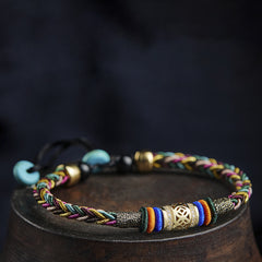 Tibetan Five-Color Bracelet – braided multicolor cord with gold tube and gemstone beads on wood base, ethnic accessory