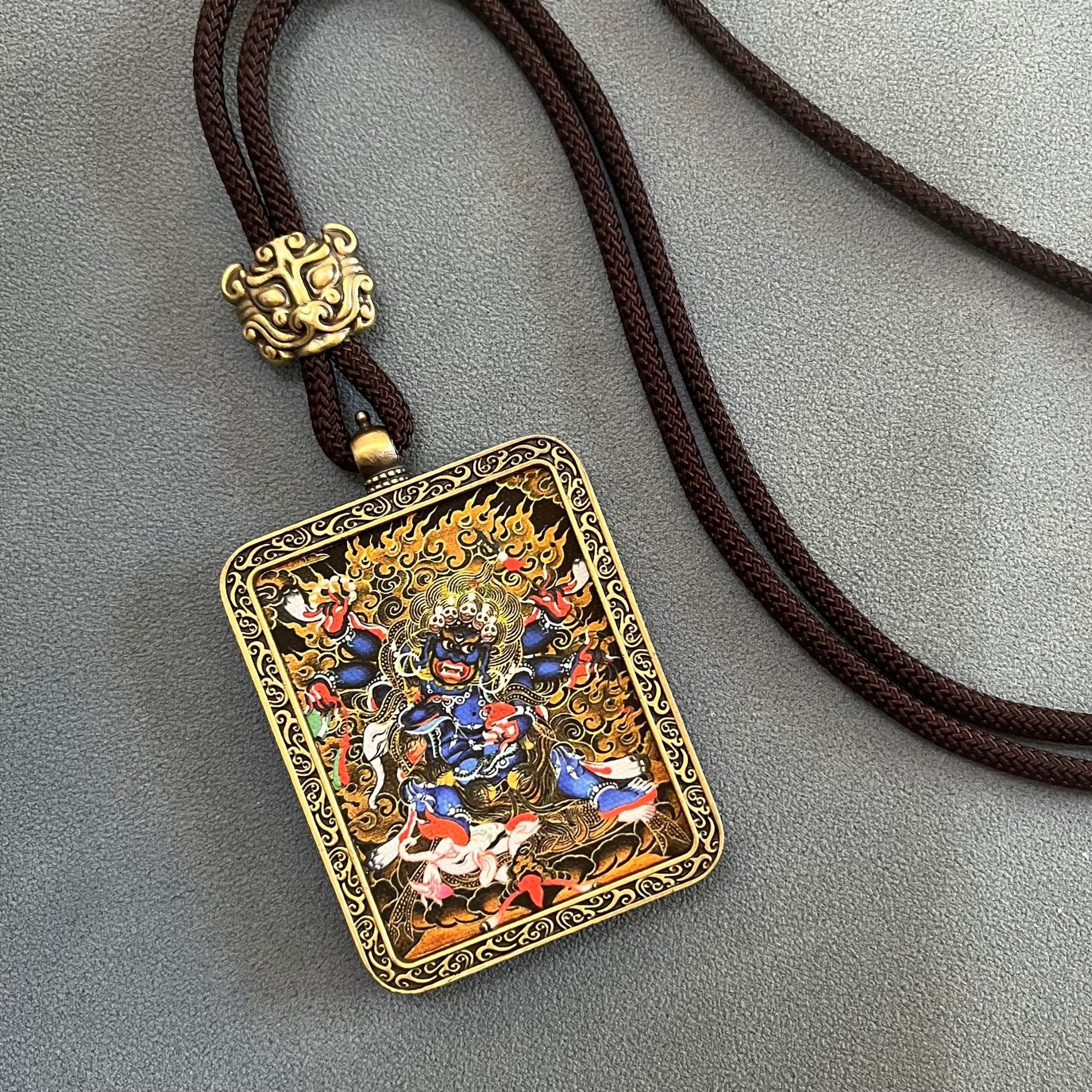Tibetan Thangka Pendant – gold-framed blue deity amulet on brown cord, grey background, Buddhist protection jewelry