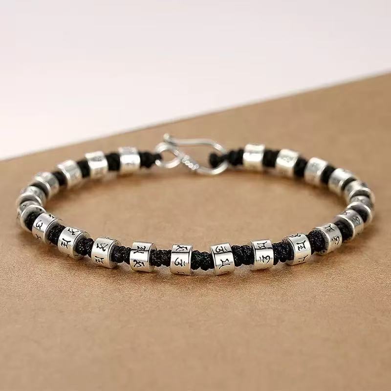 Zen Mantra Cord Bracelet – close-up of silver six-word mantra beads on black cord with metal hook closure, tan textured surface, Buddhist accessory