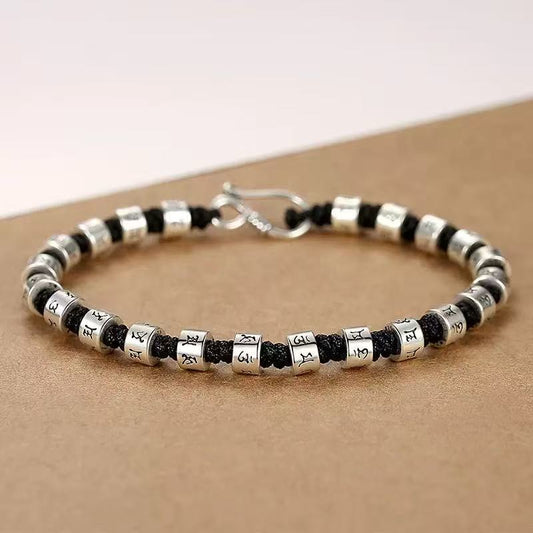 Zen Mantra Cord Bracelet – close-up of silver six-word mantra beads on black cord with metal hook closure, tan textured surface, Buddhist accessory
