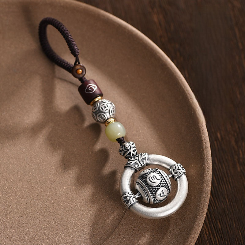 Mantra Keychain – ornate silver amulet resting on ceramic dish, jade bead detail, carved mantra