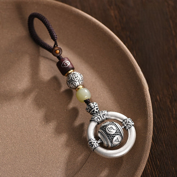 Mantra Keychain – ornate silver amulet resting on ceramic dish, jade bead detail, carved mantra