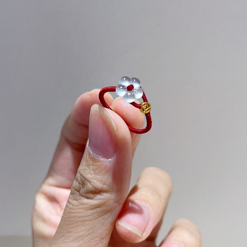 Red String Flower Ring – hand holding red cord ring with white jade flower and gold coin charm, gray background, benming year jewelry