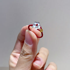 Red String Flower Ring – hand holding red cord ring with white jade flower and gold coin charm, gray background, benming year jewelry
