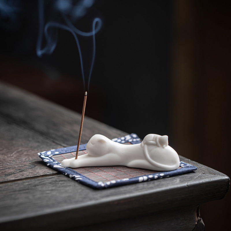 White ceramic cat incense holder stretching pose on blue floral mat, smoke