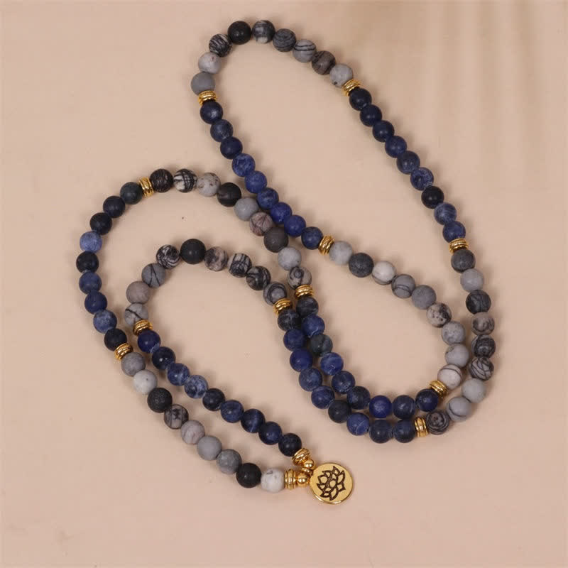 Long sodalite zebra jasper beaded necklace with navy blue web-patterned gray and charcoal beads gold-tone disc spacers and lotus flower charm in S-curve drape on beige background