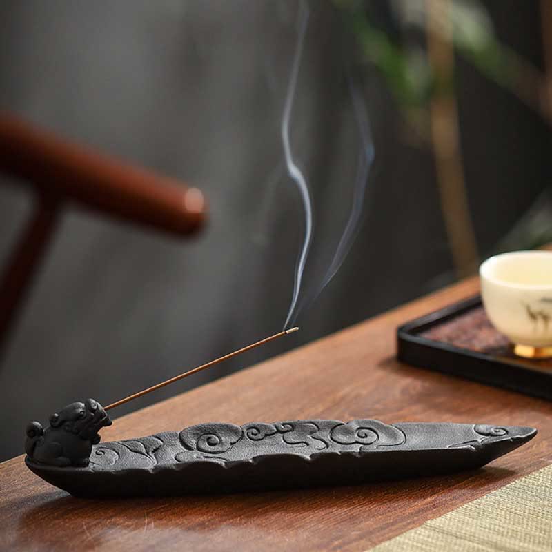 Side view of Pixiu incense holder with smoke trail, blurred teacup and fan in background, olive mat – stick incense burner