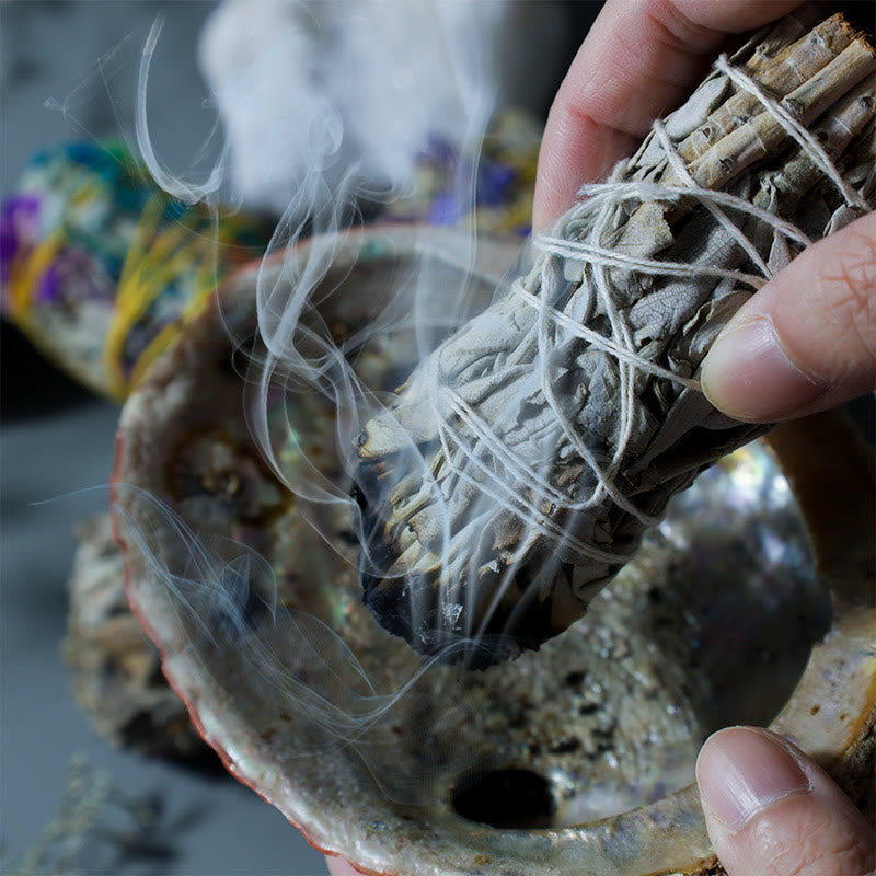 Hand holding burning sage smudge stick with smoke over iridescent abalone shell