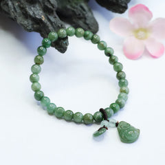 Cyan jade beaded stretch bracelet with carved Laughing Buddha figure and jade disc charm, white background with driftwood and pink flower prop accents
