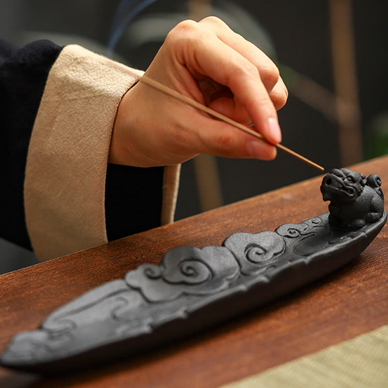 Hand placing incense stick into Pixiu beast mouth on carved cloud holder, cream cuff, warm wood – ceramic stick incense burner