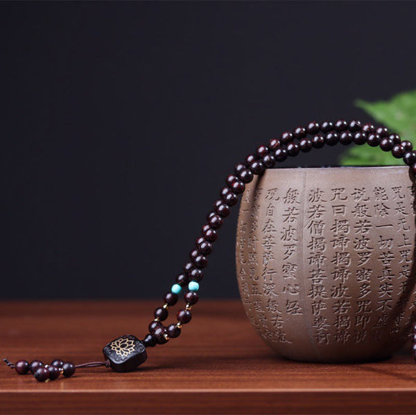 Red sandalwood mala bracelet multi-wrapped on wrist with carved dark wood guru bead featuring gold lotus inlay and scrollwork border against solid black background