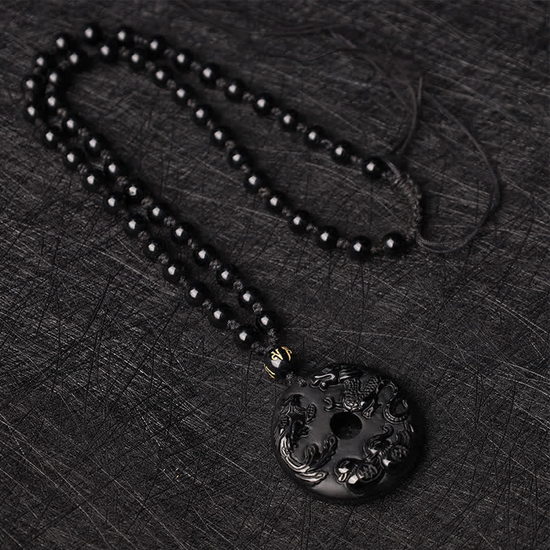 Black Obsidian Dragon Phoenix Pendant - Flat-lay beaded necklace with carved obsidian disc on dark textured fabric, Luck