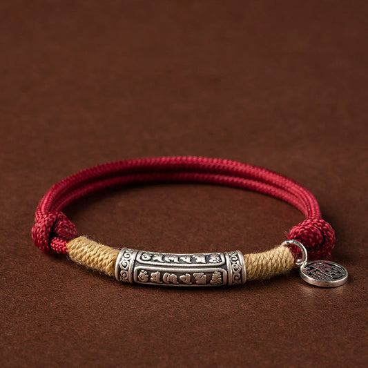 Burgundy cord bracelet with antique silver Tibetan mantra cylinder bead, natural hemp wrapping and lucky fortune coin charm on brown leather background
