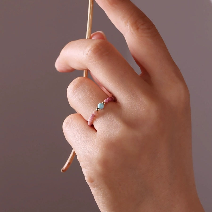 Gemstone Bead Ring – hand wearing pink beaded ring with turquoise stone, holding gold chain, gray background, natural gemstone