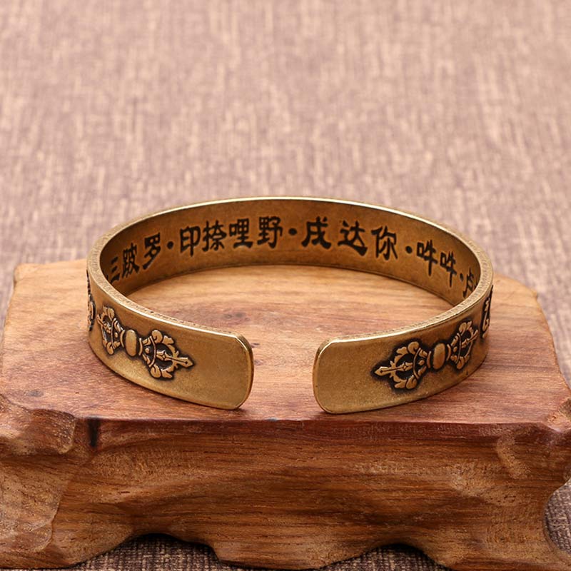Antiqued brass cuff bracelet interior showing engraved Chinese Buddhist mantra text and ornate floral terminals on wooden display stand