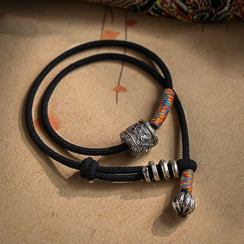 Tibetan Om Mani Padme Hum Wrap Bracelet – Black braided cord with S925 silver engraved cylinder bead, red blue yellow woven segments, beige fabric flat lay