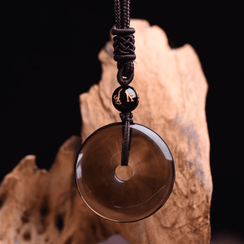 Semi-translucent dark obsidian peace buckle necklace with gold Sanskrit inscribed accent bead on black braided cord displayed on natural driftwood prop