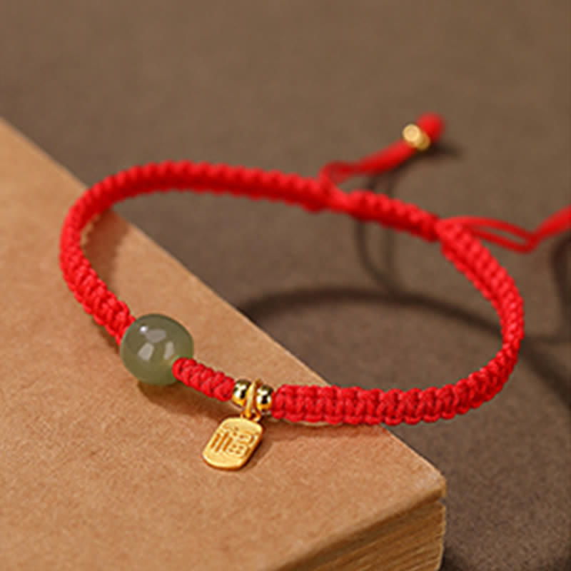 Red cord bracelet with Hetian jade green bead rectangular gold charm with Chinese fu blessing character on vintage aged book display with warm brown heritage styling