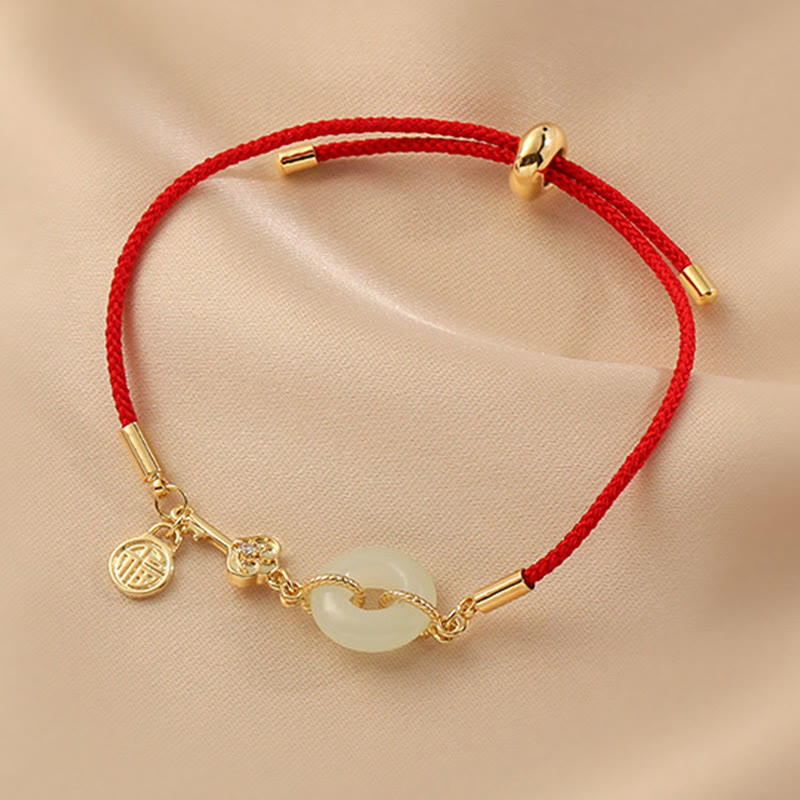 Chinese lucky red string bracelet with white jade ping an kou peace buckle, gold fortune character charm and crystal accent on champagne fabric background