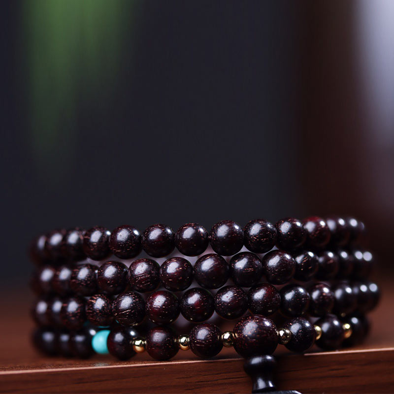 108 bead red sandalwood mala bracelet coiled with turquoise accent and gold lotus guru bead on dark surface with blurred green foliage background