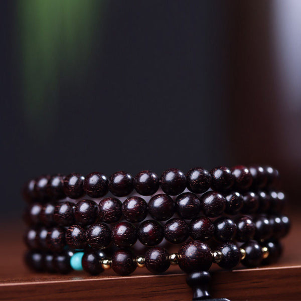 108 bead red sandalwood mala bracelet coiled with turquoise accent and gold lotus guru bead on dark surface with blurred green foliage background