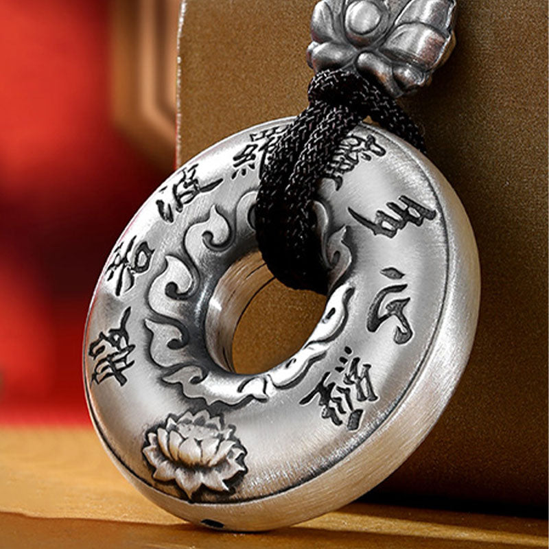 Silver Heart Sutra Pendant – close-up silver disc with relief lotus and flame motifs on black cord, warm bokeh background