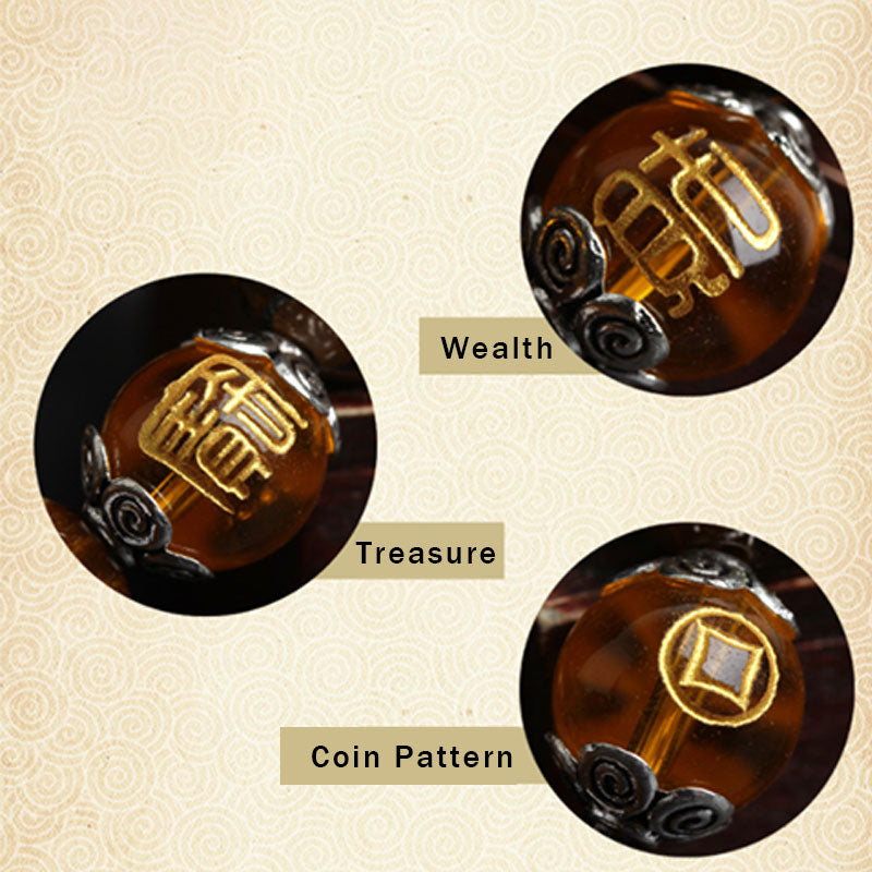 Detail close-up of three carved amber cylindrical beads with gold Wealth character, Treasure character and coin pattern inlays, silver scroll caps on cream Chinese cloud pattern background