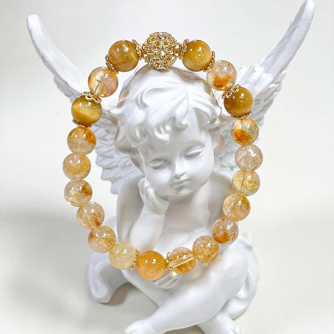 Natural Citrine Bracelet – golden yellow and clear citrine beads with gold filigree spacer beads, draped over white angel figurine wings, clean white background, artistic product display
