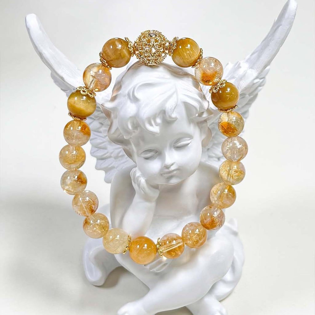 Natural Citrine Bracelet – golden yellow and clear citrine beads with gold filigree spacer beads, draped over white angel figurine wings, clean white background, artistic product display