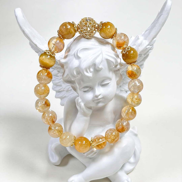 Natural Citrine Bracelet – golden yellow and clear citrine beads with gold filigree spacer beads, draped over white angel figurine wings, clean white background, artistic product display