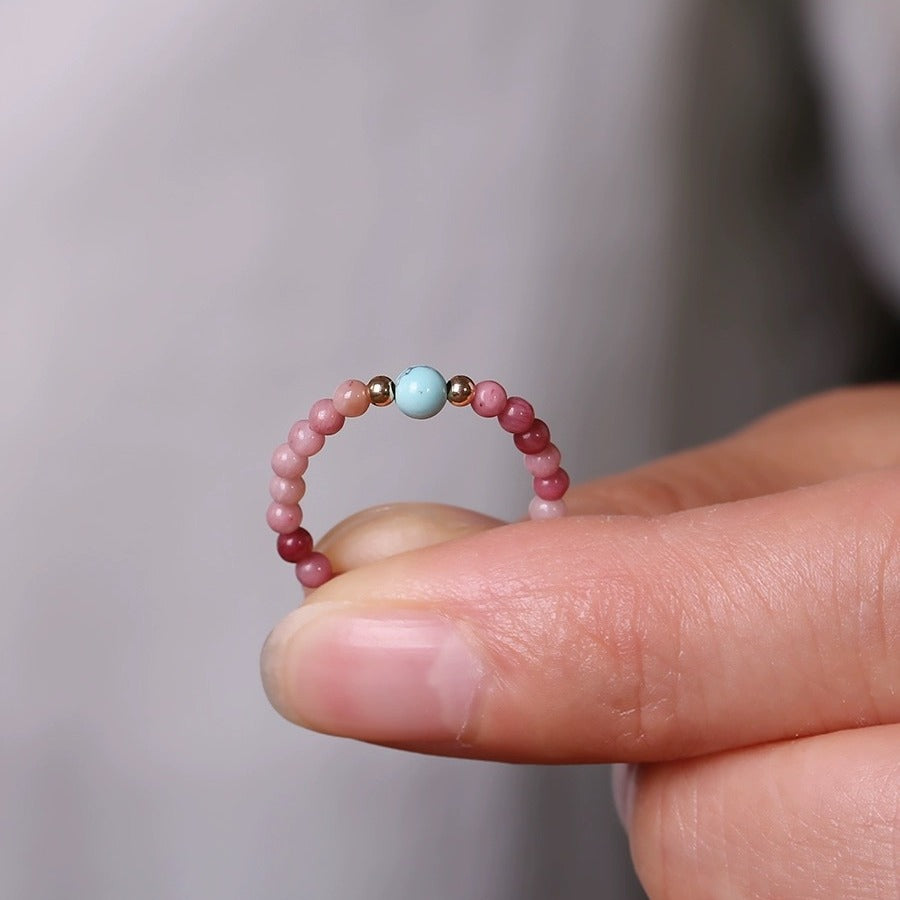 Gemstone Bead Ring – fingers holding delicate pink bead ring, blue turquoise center, gold accents, soft gray bokeh background