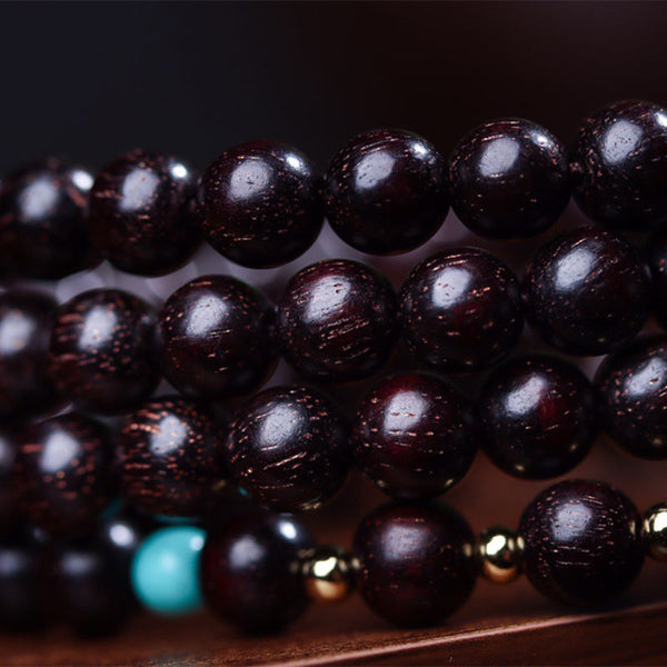 Macro close-up of polished red sandalwood mala beads showing natural wood grain, turquoise marker bead and gold-toned metal spacers on dark wood surface