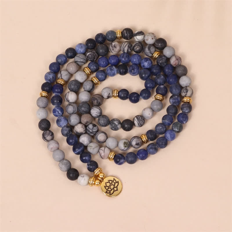 108 bead mala bracelet necklace with sodalite howlite and onyx beads in blue gray and black gold textured spacer beads and circular lotus flower pendant on warm beige backdrop