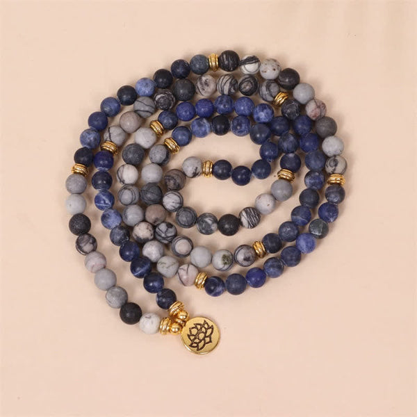 108 bead mala bracelet necklace with sodalite howlite and onyx beads in blue gray and black gold textured spacer beads and circular lotus flower pendant on warm beige backdrop